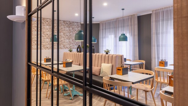 A modern dining area with wooden tables and chairs, a cushioned seating area, and pendant lighting. - Garner Hotel Mannheim City by IHG (Mannheim)