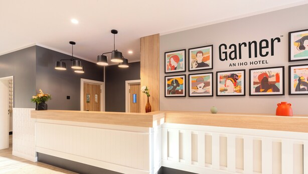 A hotel reception area with a modern design, featuring a reception desk, framed artwork on the wall, and a sign that reads "Garner AN IHG HOTEL".