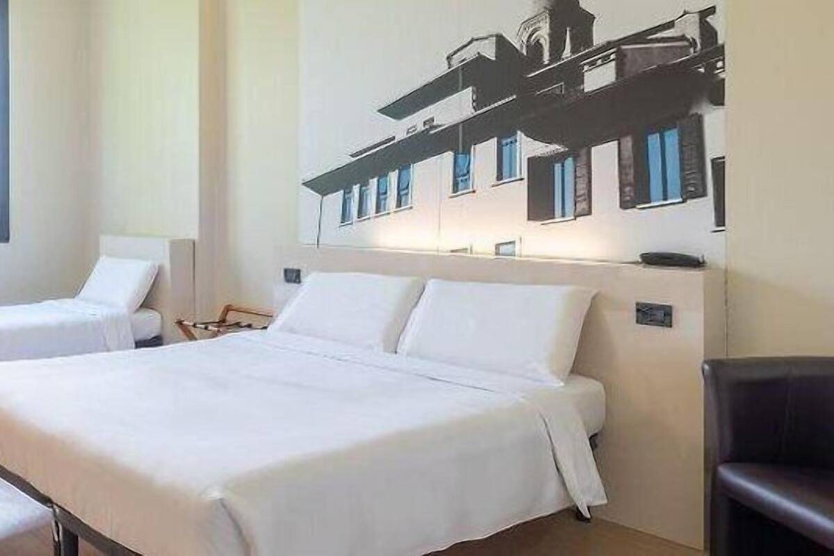 A hotel room with two beds, a window with curtains, and a wall-mounted artwork of a building.