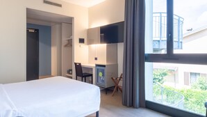 Habitación de hotel con cama, escritorio, silla, televisión y ventana con vistas a zonas verdes y edificios.
