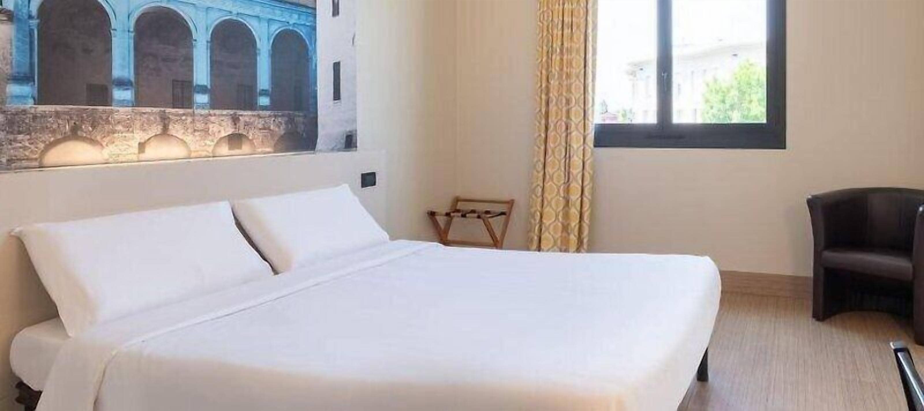 A hotel room with a large bed, a small desk, a chair, and a window with curtains.