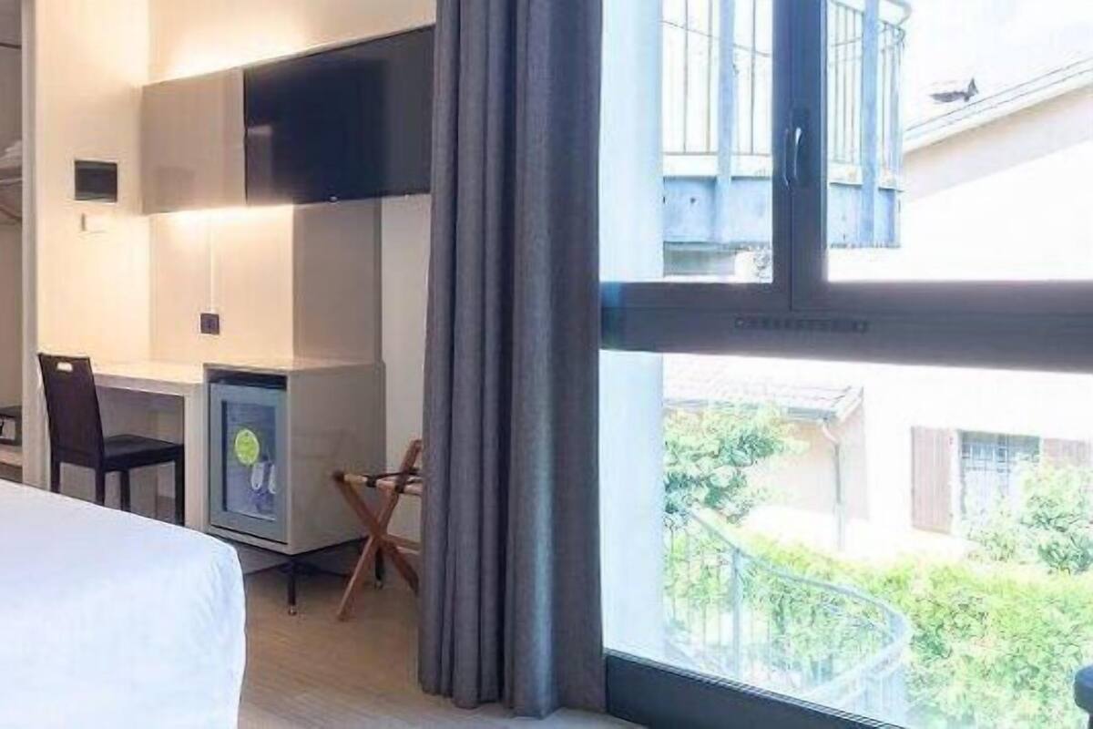 A hotel room with a bed, a desk with a chair, a TV, and a window with a view of greenery outside.