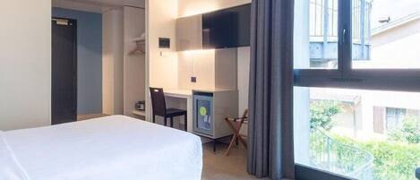 A hotel room with a bed, a desk with a chair, a TV, and a window with a view of greenery outside.