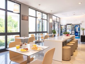 A modern dining area with large windows, a central island with seating, and a visible shop in the background. - B&B Hotel Mantova (San Giorgio di Mantova)