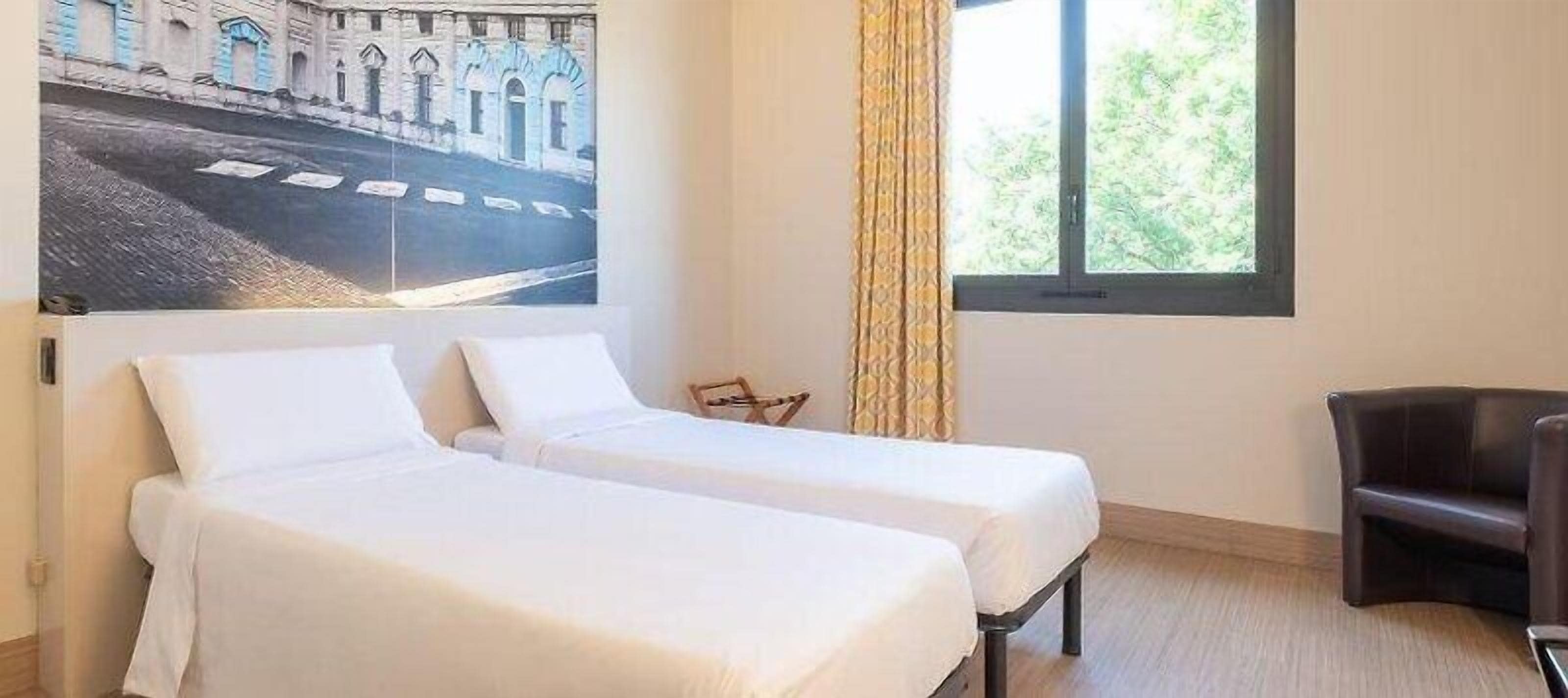 Une chambre d’hôtel avec deux lits simples, une fenêtre avec des rideaux, une chaise et une œuvre d’art accrochée au mur.