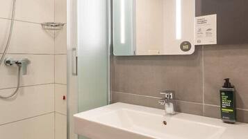 A bathroom with a shower, a sink, and a mirror. There is a soap dispenser on the sink and a sign on the wall.