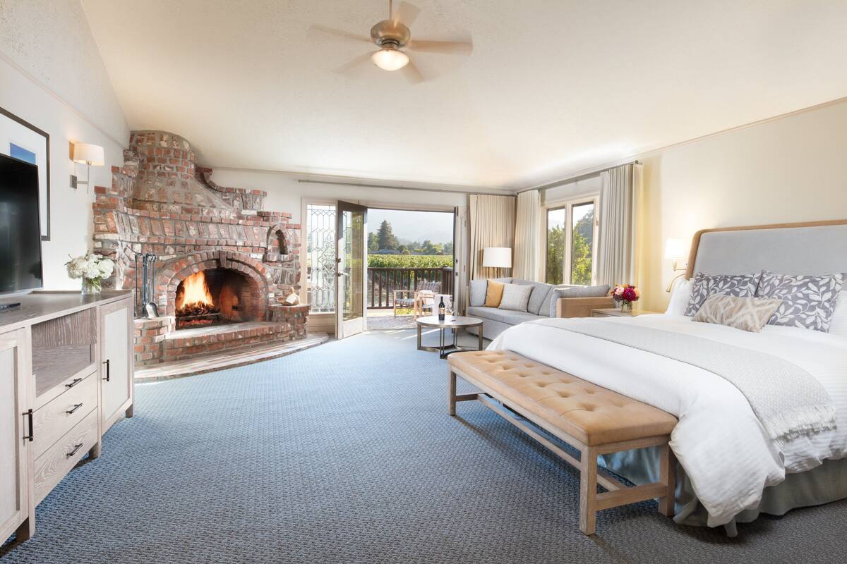 A bedroom with a fireplace, a bed, a bench, a TV, and a balcony with a view.