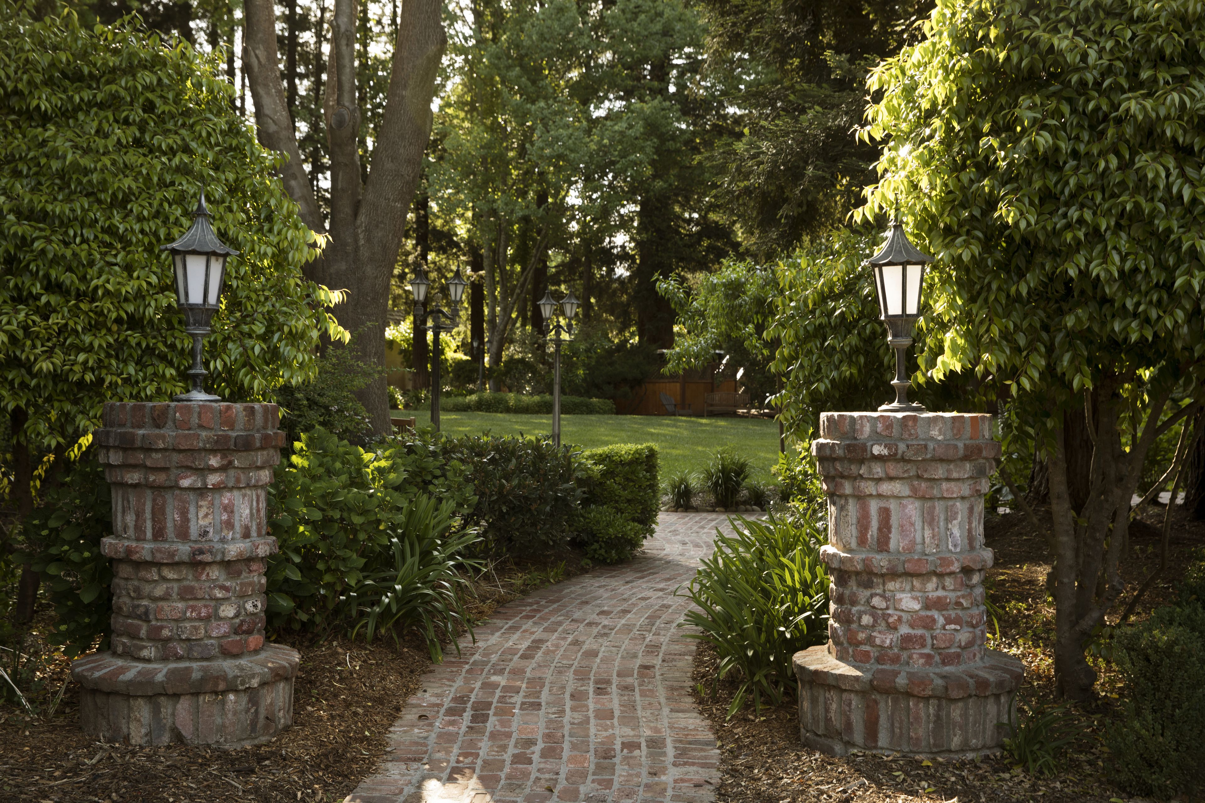 A brick pathway flanked by two lantern-style street lamps, surrounded by lush greenery and trees.
