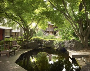 A serene garden with a pond, wooden benches, and a stone pathway leading to a building with a balcony.