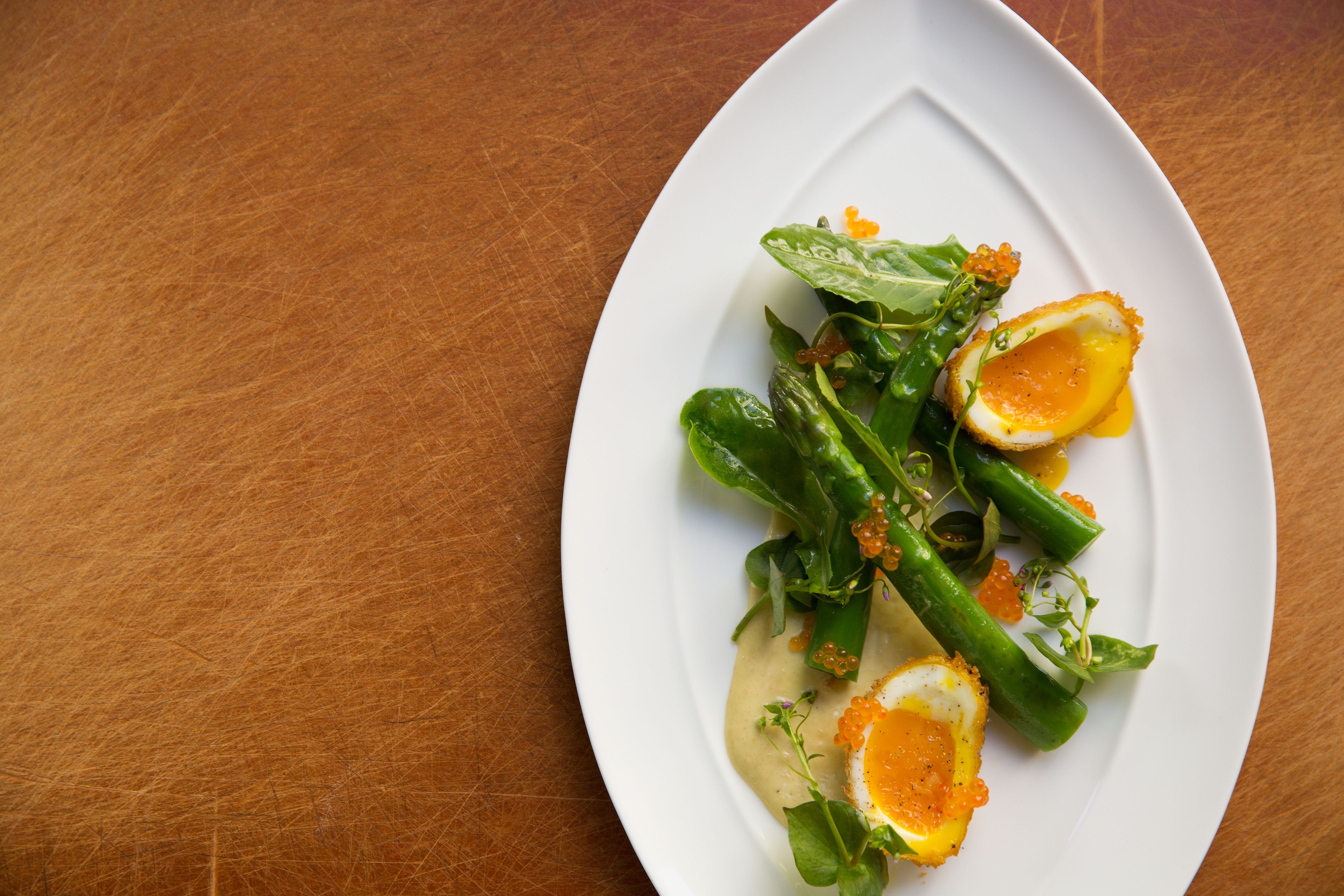 Een witte bord met een salade van bladgroenten, asperges en een gepocheerd ei op een houten tafel.