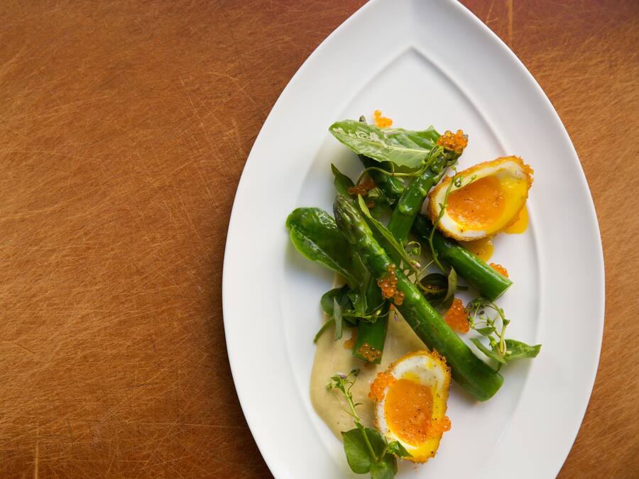 Een witte bord met een salade van bladgroenten, asperges en een gepocheerd ei op een houten tafel.