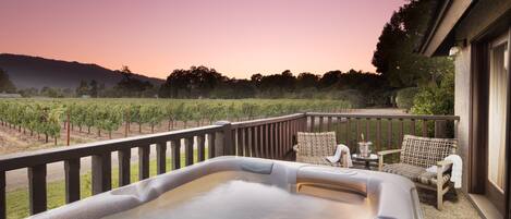 A hot tub on a balcony with a vineyard view, wicker chairs, and a small table with a drink.
