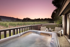 A hot tub on a balcony with a vineyard view, wicker chairs, and a small table with a drink.