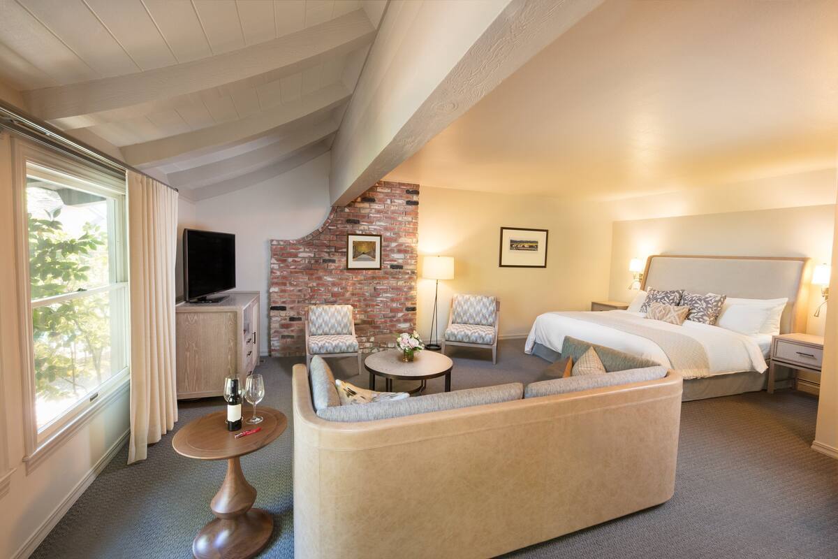 A hotel room with a bed, two armchairs, a sofa, a TV, a lamp, and a small table with a bottle and glass.