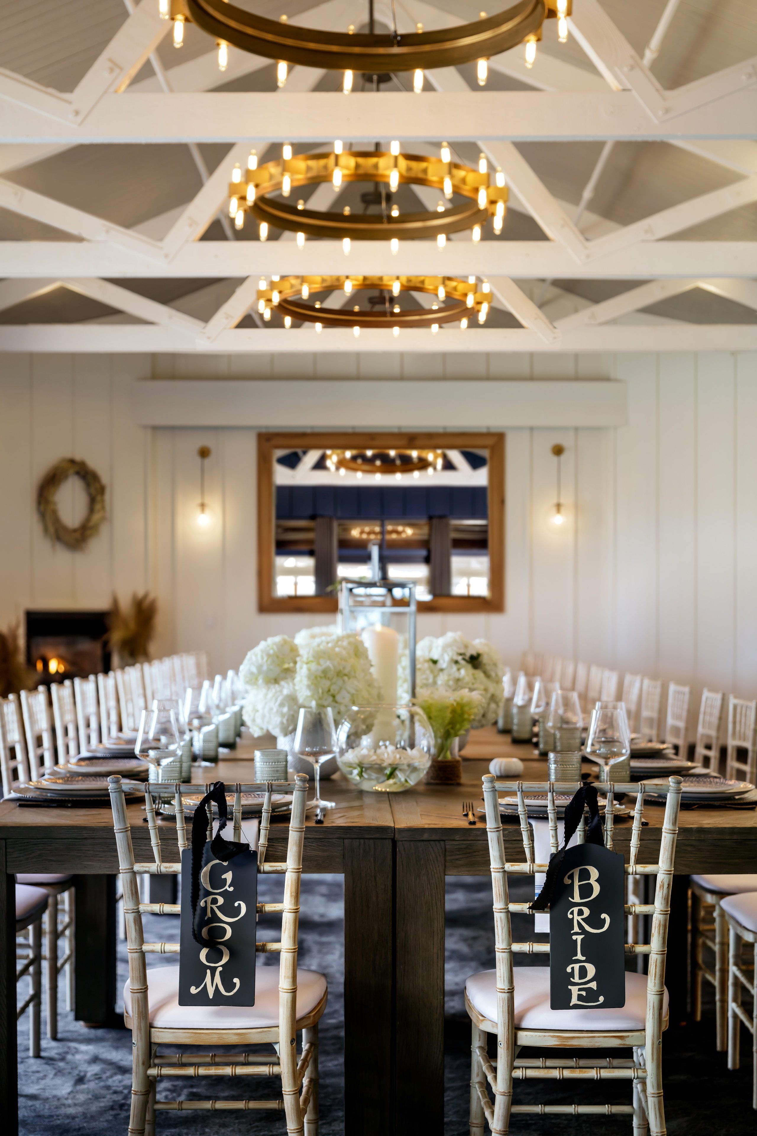 Een feestzaal met een lange tafel gedekt voor een bruiloftsreceptie, met witte stoelen voorzien van borden met 'Groom' en 'Bride', een kroonluchter en een groot bloemenarrangement.