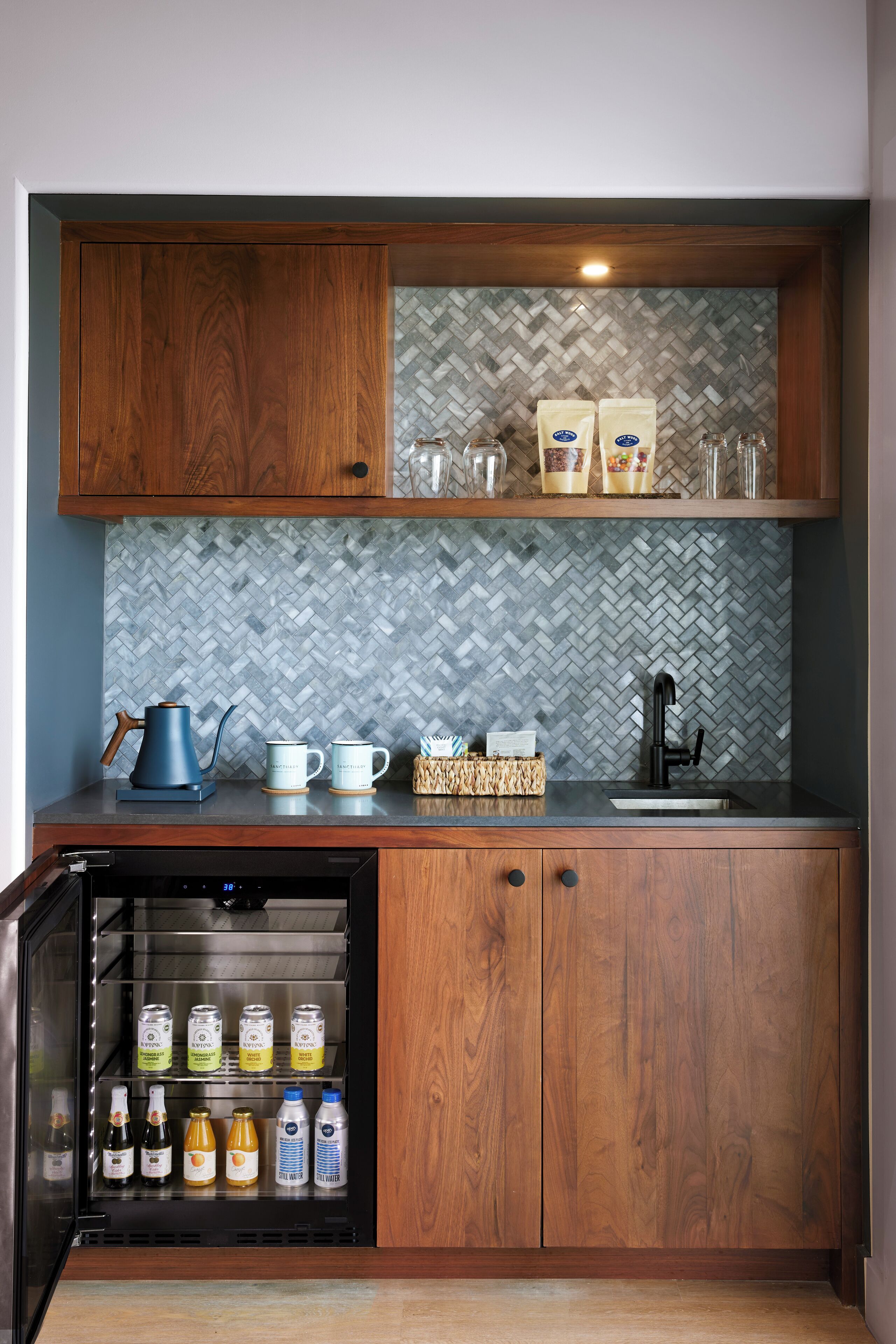 Een keuken met houten kastjes, een backsplash van mozaïektegels in een visgraatpatroon en een inbouwkoelkast vol drankjes.