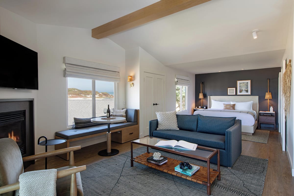 Quarto de hotel moderno com uma cama grande, um sofá, uma mesa pequena, uma lareira e vista para o exterior.