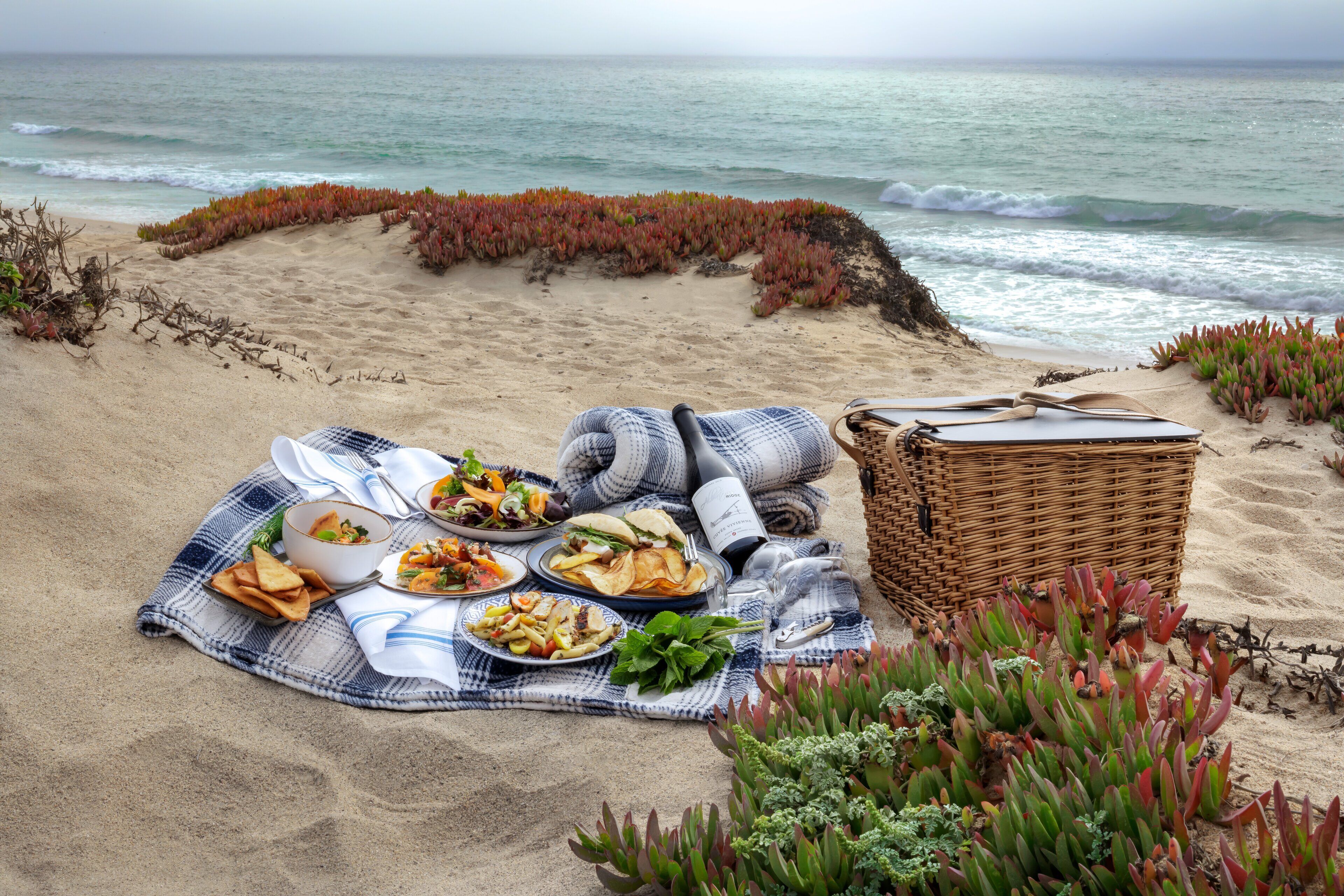 A beach picnic setup with food, a basket, and a blanket on the sand.