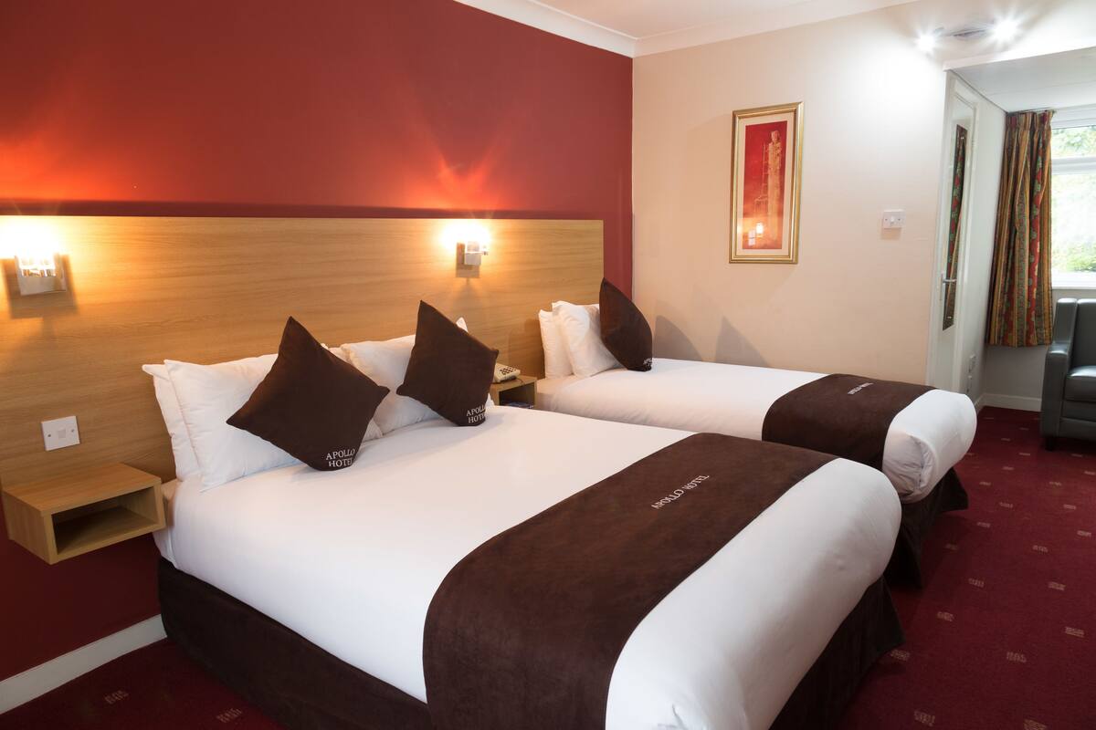 A hotel room with two beds, each with white linens and brown headboards. There are wall-mounted lights above the beds and a framed picture on the wall.