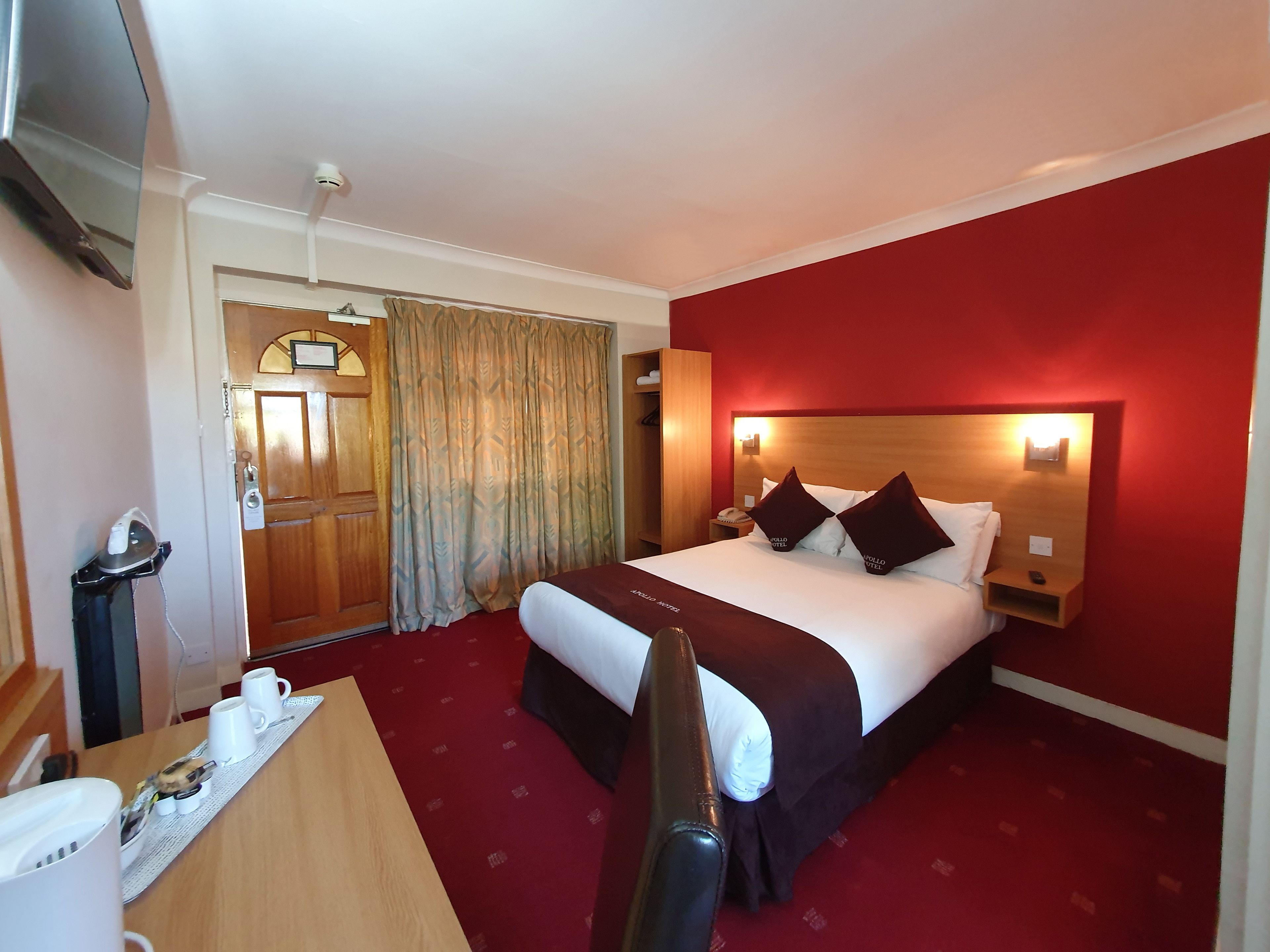 A hotel room with a bed, a desk with a telephone, a chair, a bedside table with a lamp, and a red carpeted floor.