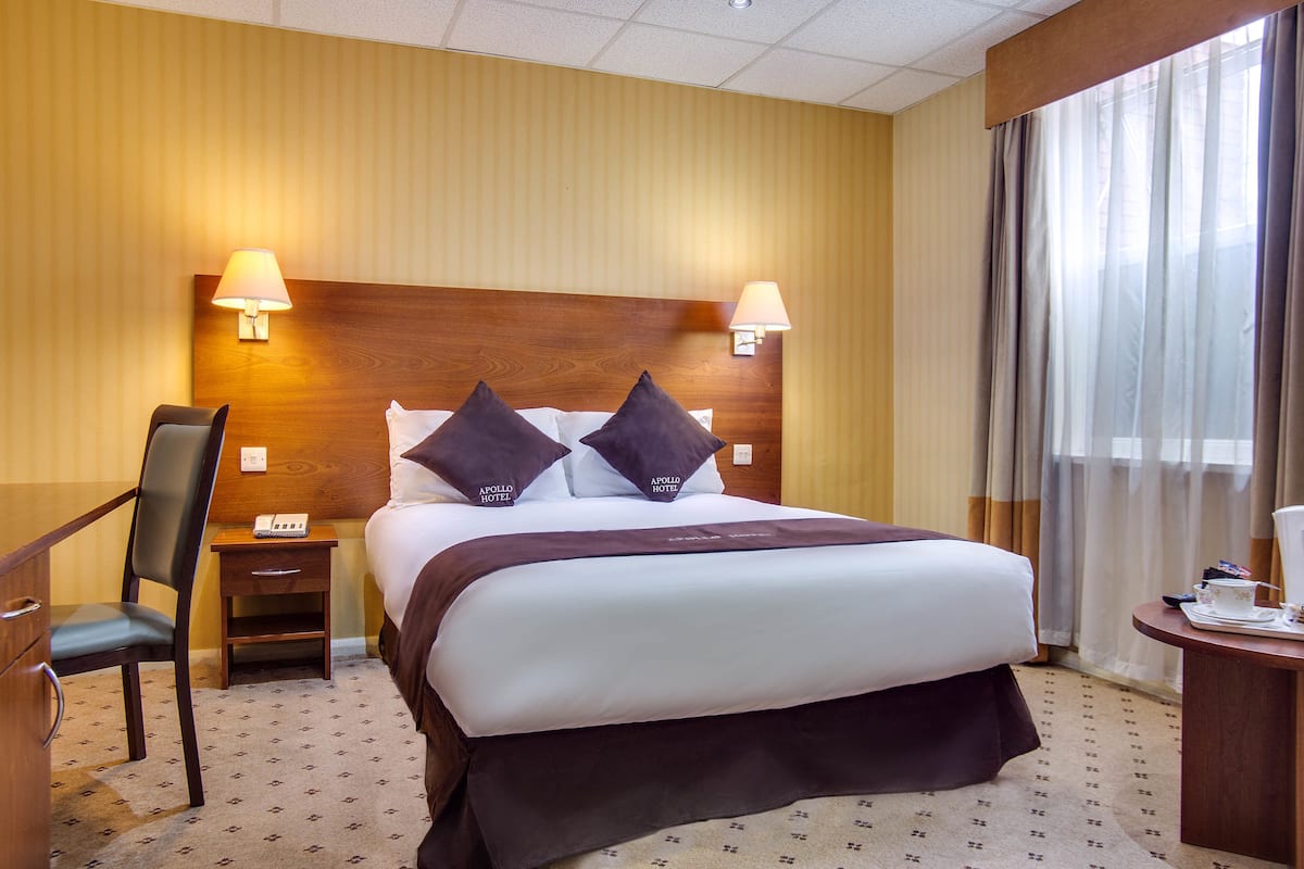 A hotel room with a large bed, two bedside lamps, a wooden headboard, a desk with a chair, and a window with curtains.