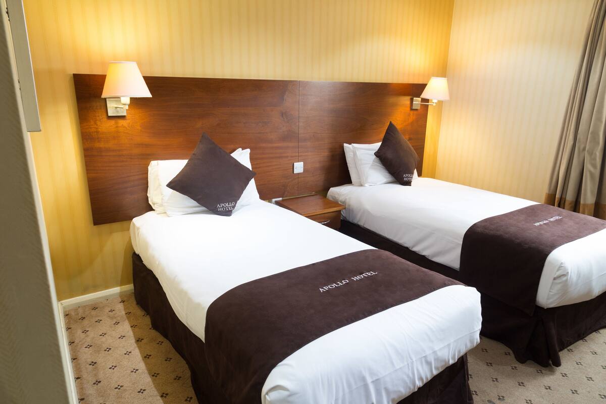 A hotel room with two beds, each with a brown headboard, white bedding, and a pillow with the hotel name. There are two wall-mounted lamps on either side of the headboard.