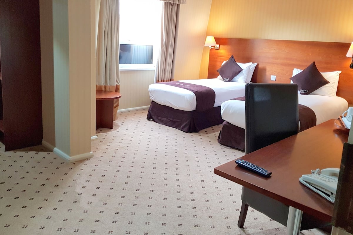 A hotel room with two beds, a desk with a phone and a remote control, a lamp, and a window with curtains.