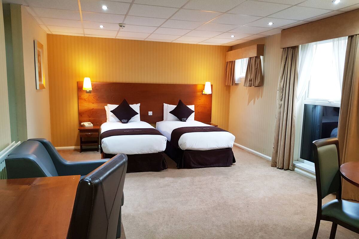 A hotel room with two beds, a desk, and a chair. There are two bedside lamps, a window with curtains, and a radiator.