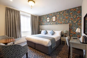 Een hotelkamer met een groot bed, een bureau, een stoel, een raam met gordijnen en bloemenbehang.