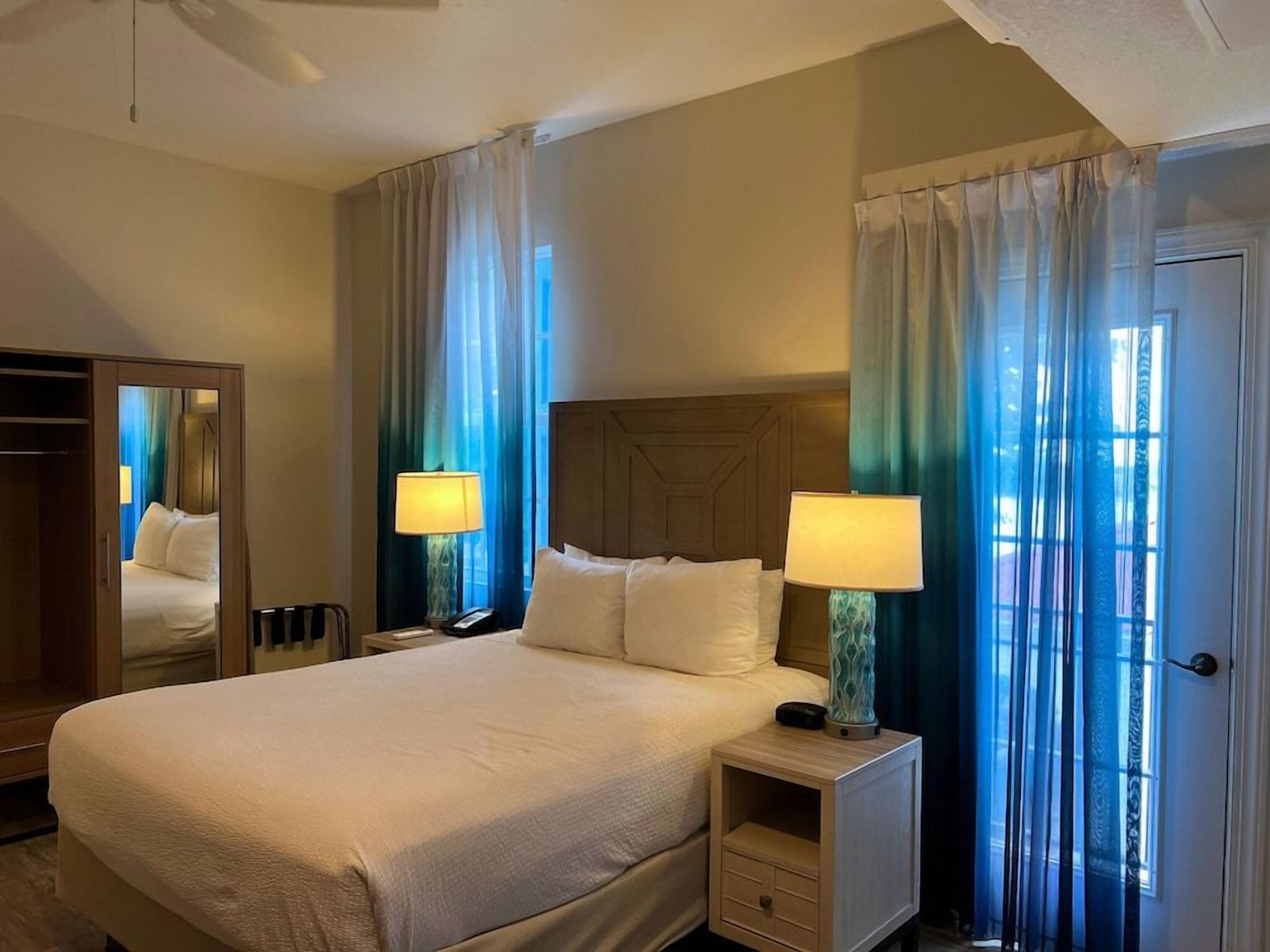A hotel room with a large bed, two bedside tables, a mirror, and a view through sheer curtains.