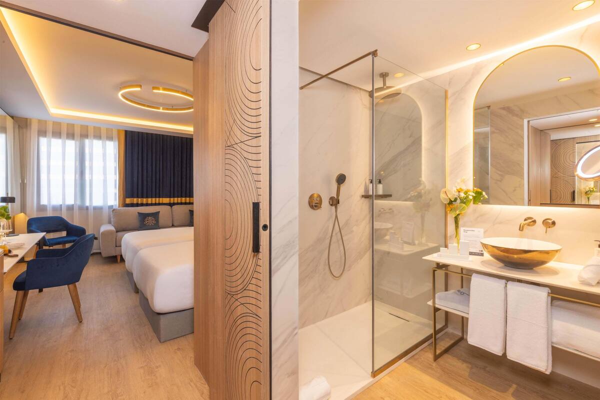 A modern hotel room with a wooden panel wall, a glass-enclosed shower, a marble bathroom, a sofa, and a dining area with a table and chairs.