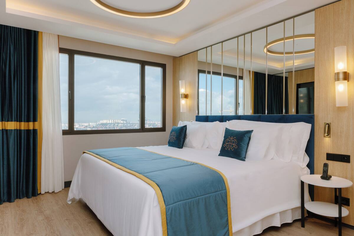 A hotel room with a large bed, a bedside table, a mirror, and a view of the city through the window.