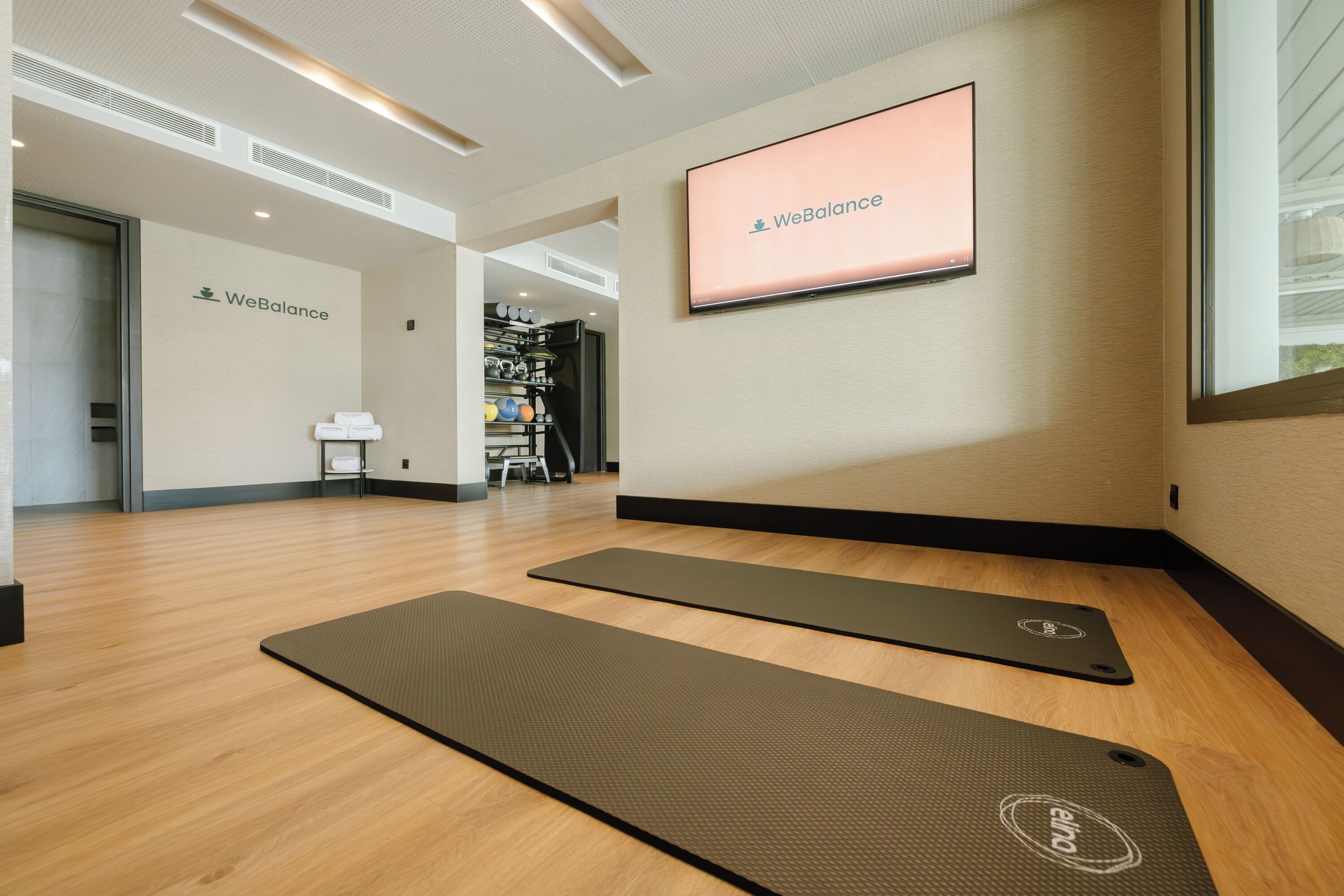 A WeBalance studio with exercise mats, a TV screen, and a view of the gym area.
