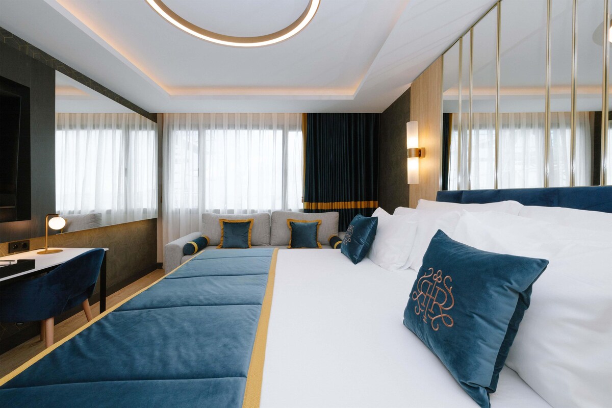 Habitación de hotel con una cama grande, un escritorio y una silla. Hay un televisor en la pared y una ventana con cortinas.
