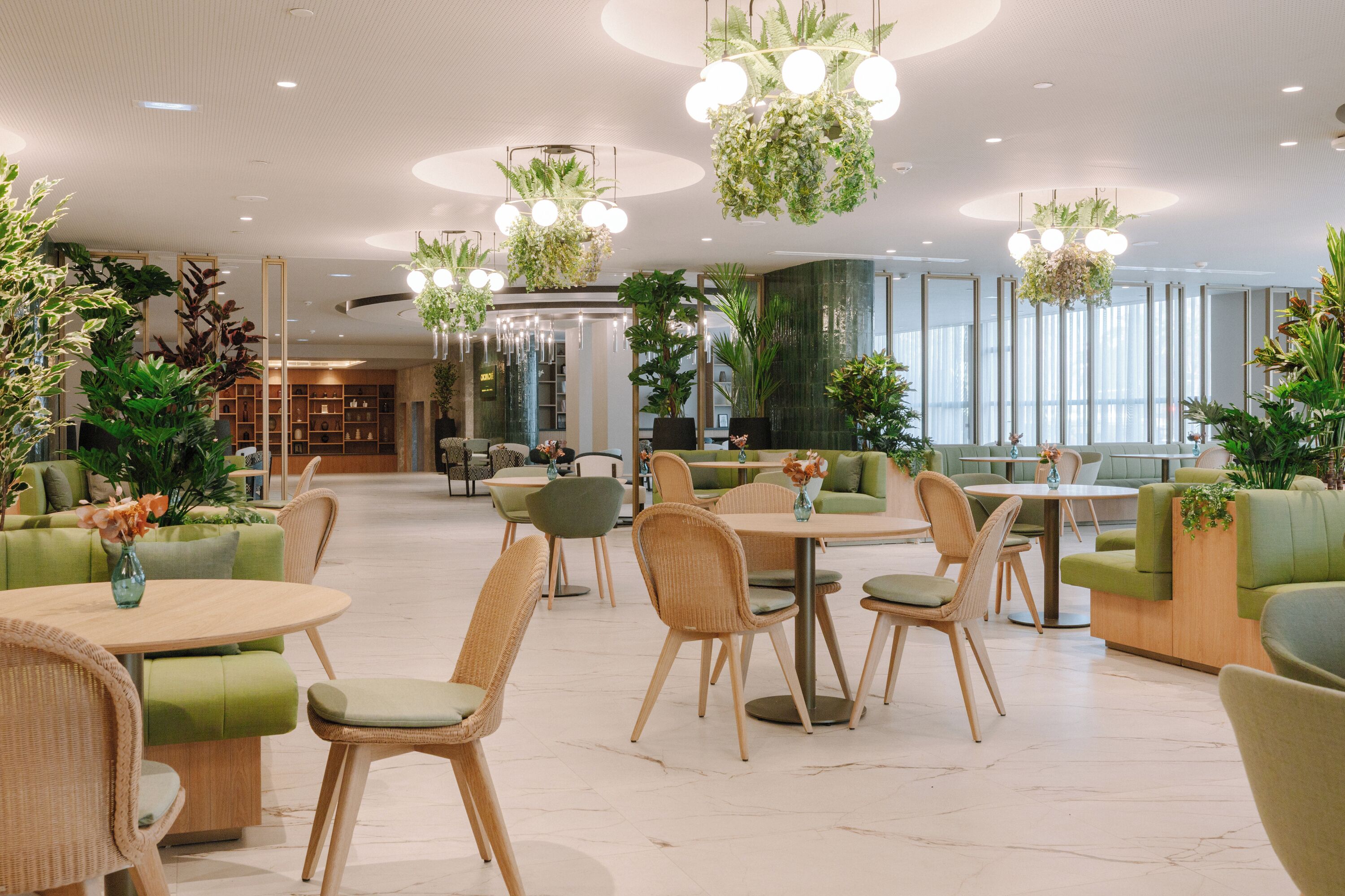 A modern hotel lobby with green seating areas, round tables, and hanging plants.