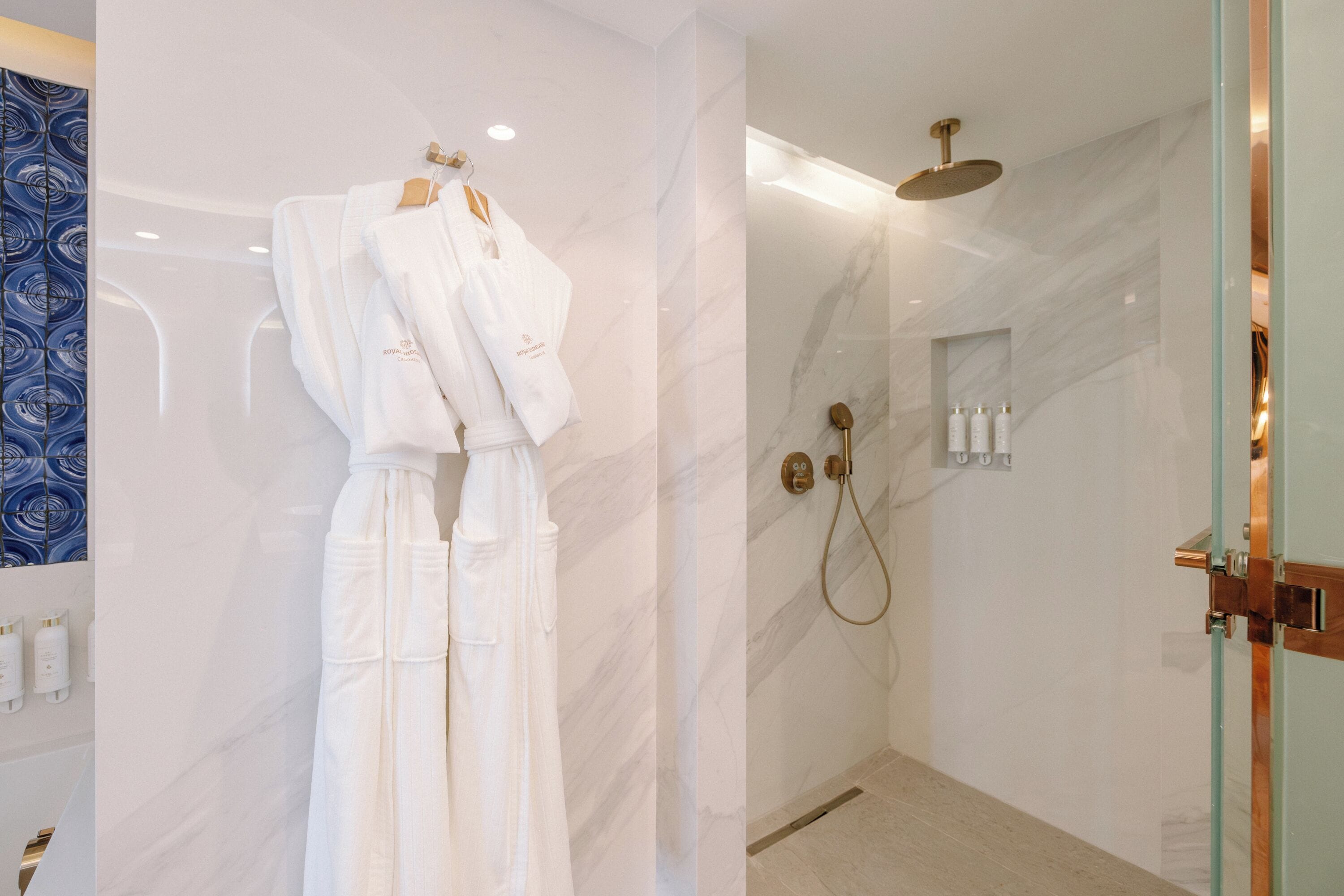 A modern bathroom with a marble wall, a shower with a handheld showerhead, and a towel rack with white robes.
