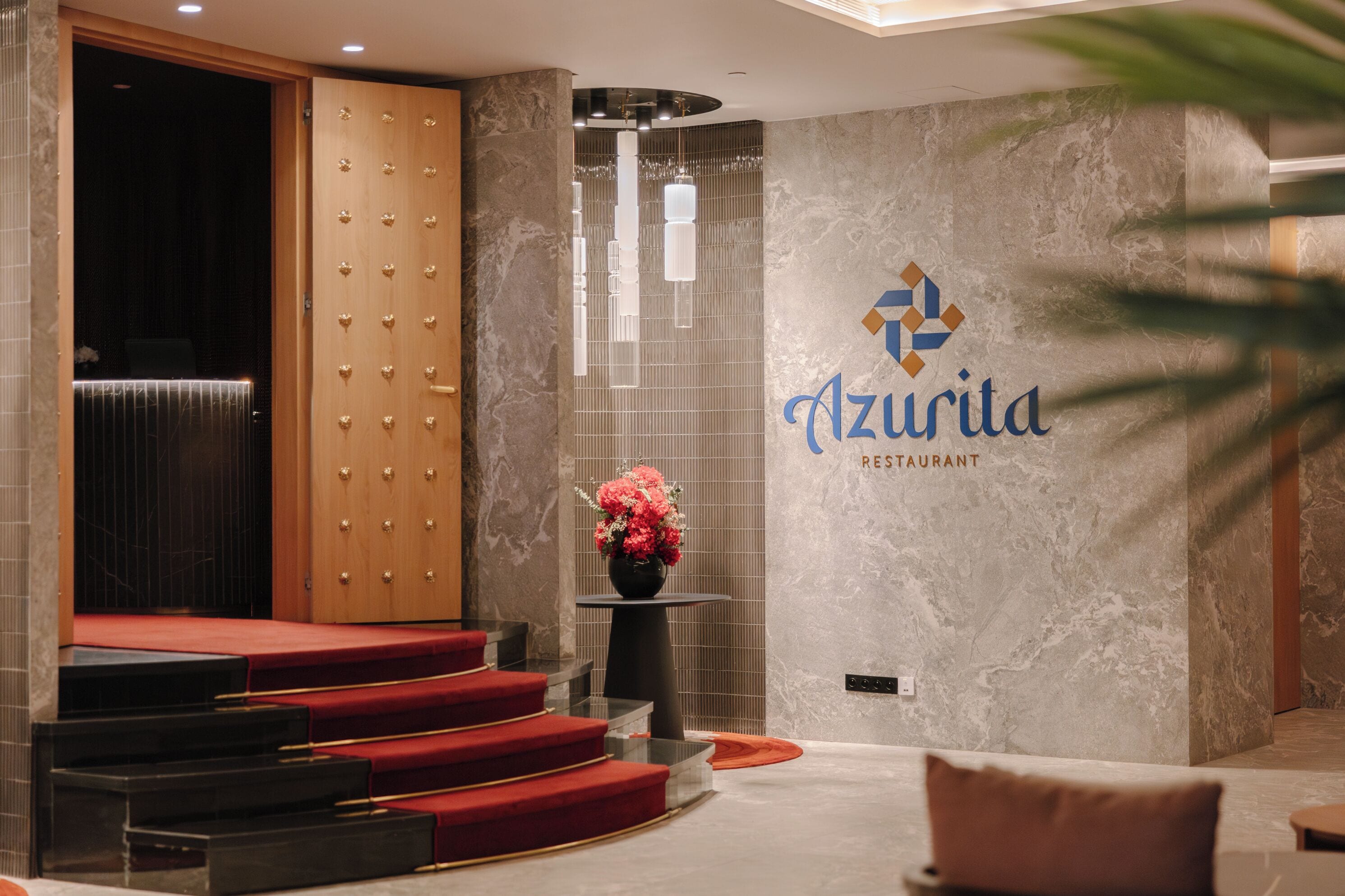 Un hall d’entrée avec un escalier recouvert d’un tapis rouge, un comptoir d’accueil et un mur sur lequel est inscrit en lettres stylisées le nom « Azurita Restaurant ».