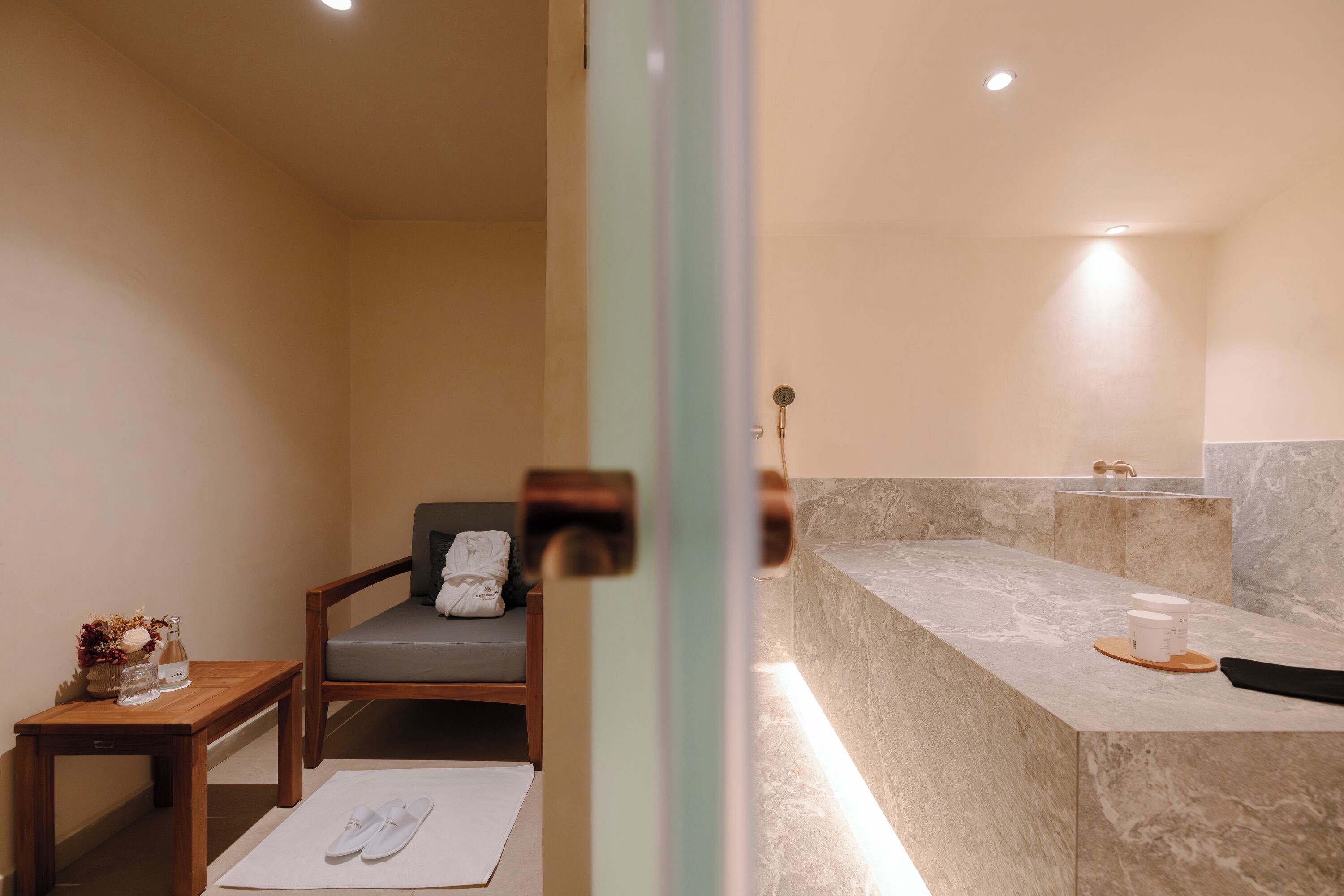 Un bagno con un lavabo in marmo, una sedia di legno con un asciugamano e un tavolino con una bottiglia e fiori.