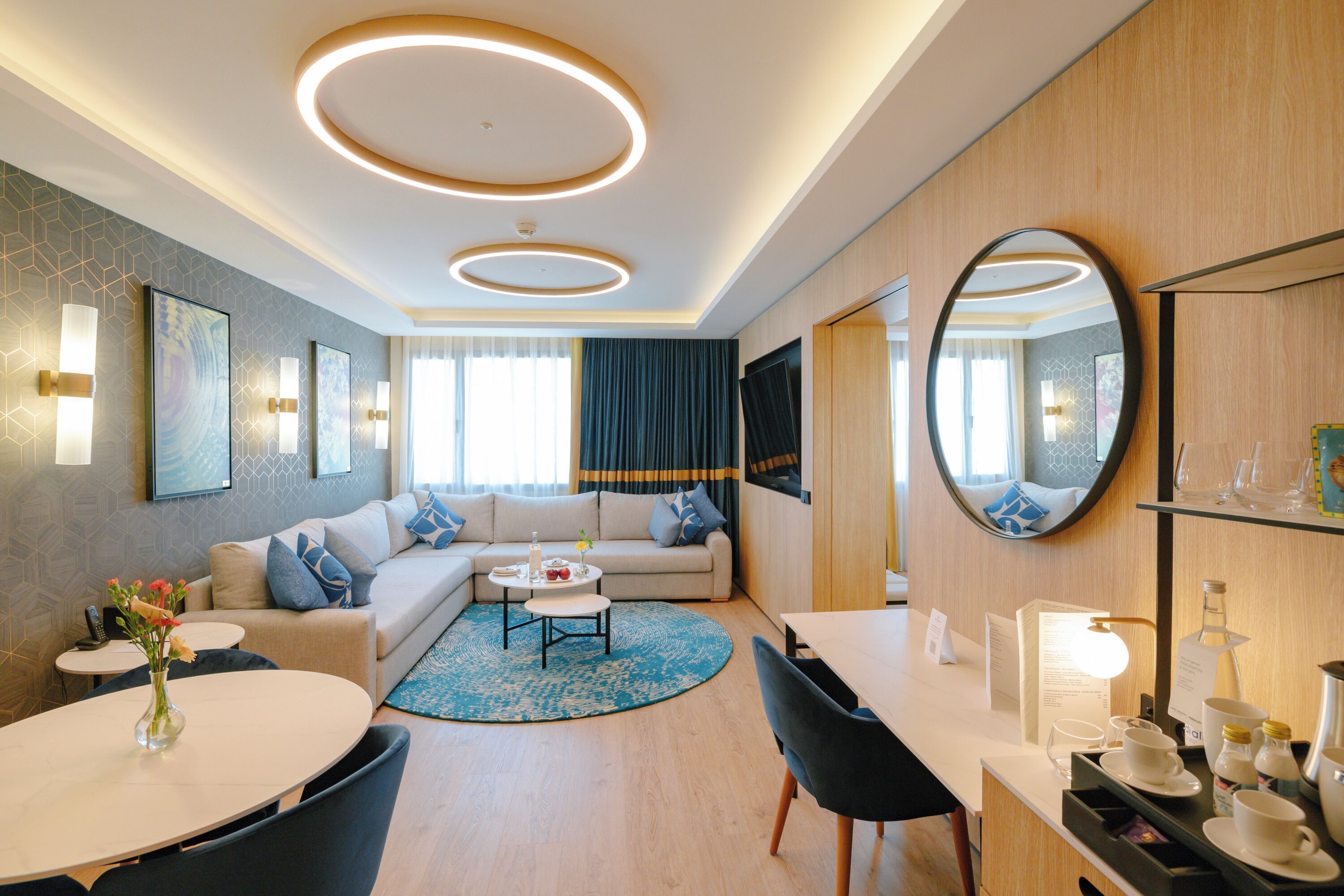 Une chambre d’hôtel moderne équipée d’un canapé, d’une table basse ronde, d’une table à manger, de chaises, d’un miroir et d’un téléviseur.