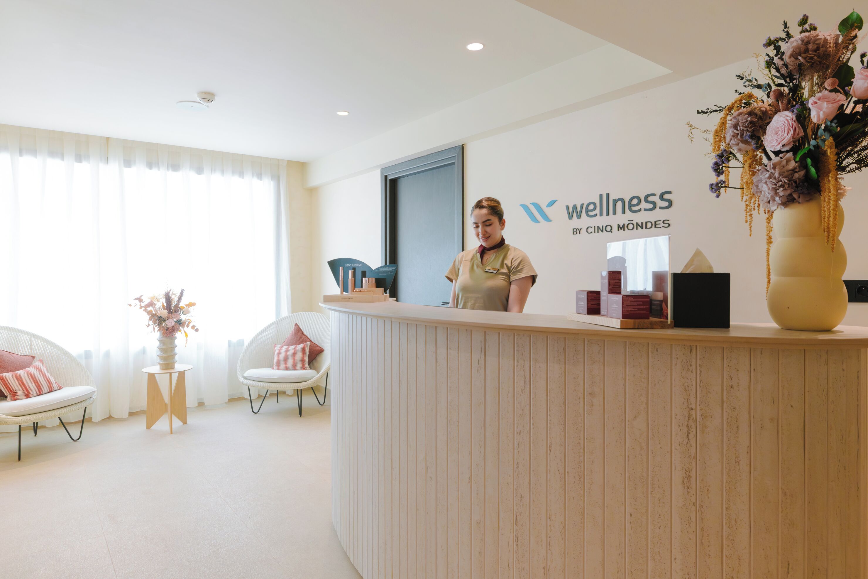 A reception desk with a logo, a receptionist, two chairs, a small table with a vase, and a large vase with flowers.
