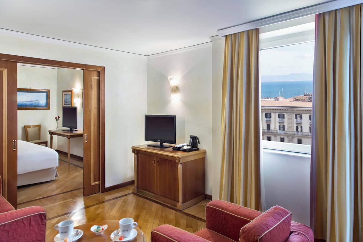 A hotel room with a bed, a TV on a wooden stand, a desk with a chair, a dining table with cups and saucers, and a view of the sea through a window with curtains.