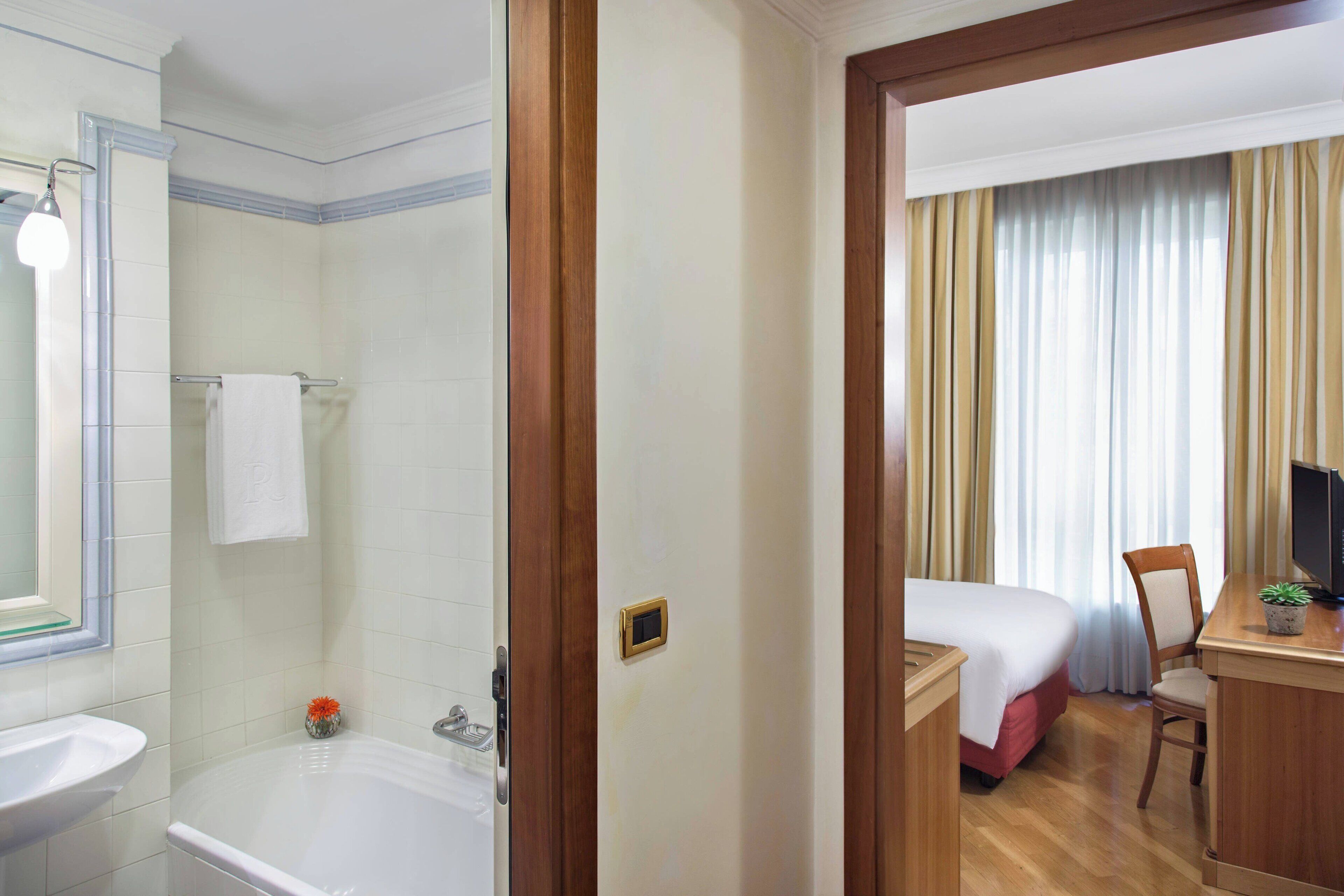 Ein Hotelzimmer mit Badewanne, einem Bett, einem Schreibtisch mit Stuhl und einem Vorhangfenster.