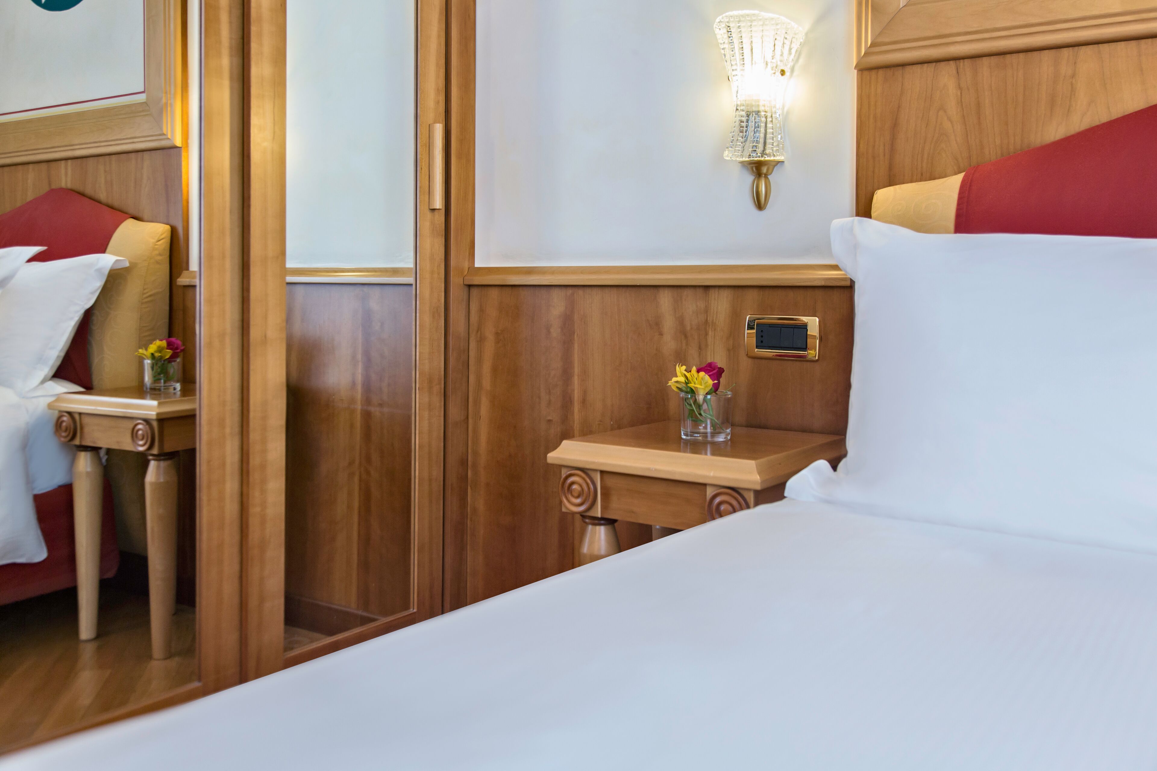 Habitación de hotel con cabecera de madera, mesita de noche con un florero y una lámpara de pared.