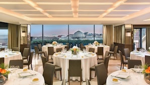 A restaurant with round tables set for dining, a cityscape view through large windows, and a modern ceiling design. - Renaissance Naples Hotel Mediterraneo (Naples)