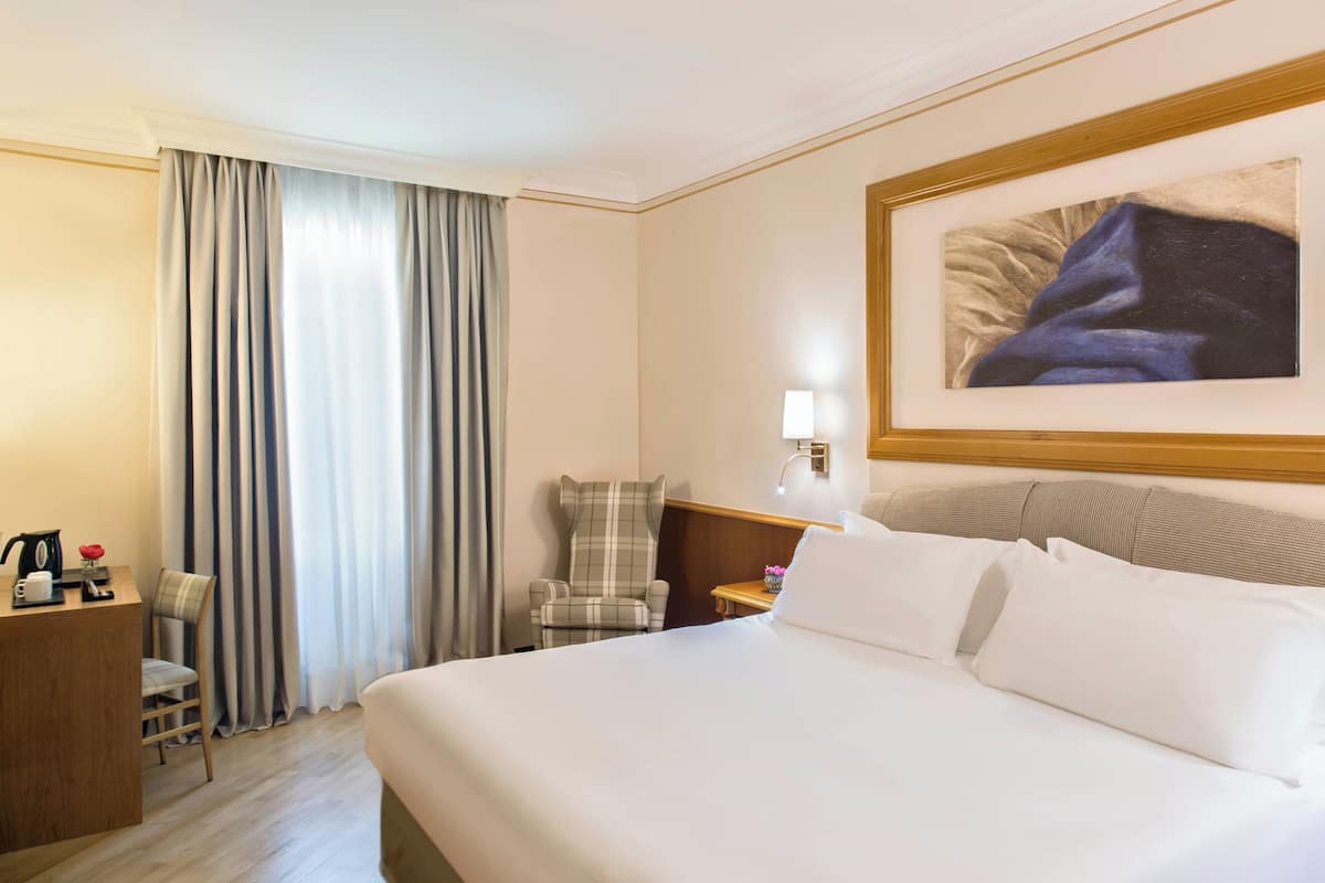 A hotel room with a large bed, a desk with a coffee maker, a chair, a nightstand with a lamp, and a painting on the wall.