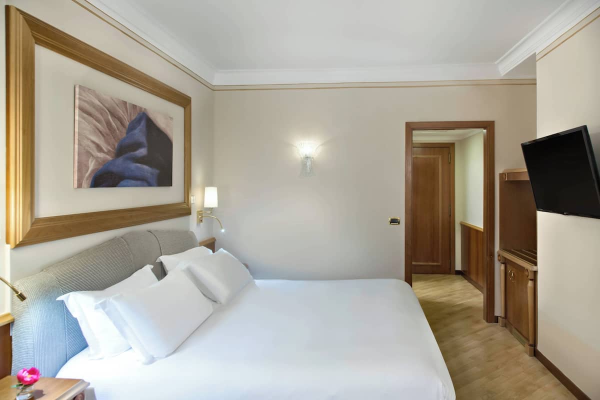 A hotel room with a large bed, a flat-screen TV, a painting on the wall, and a hallway leading to another room.