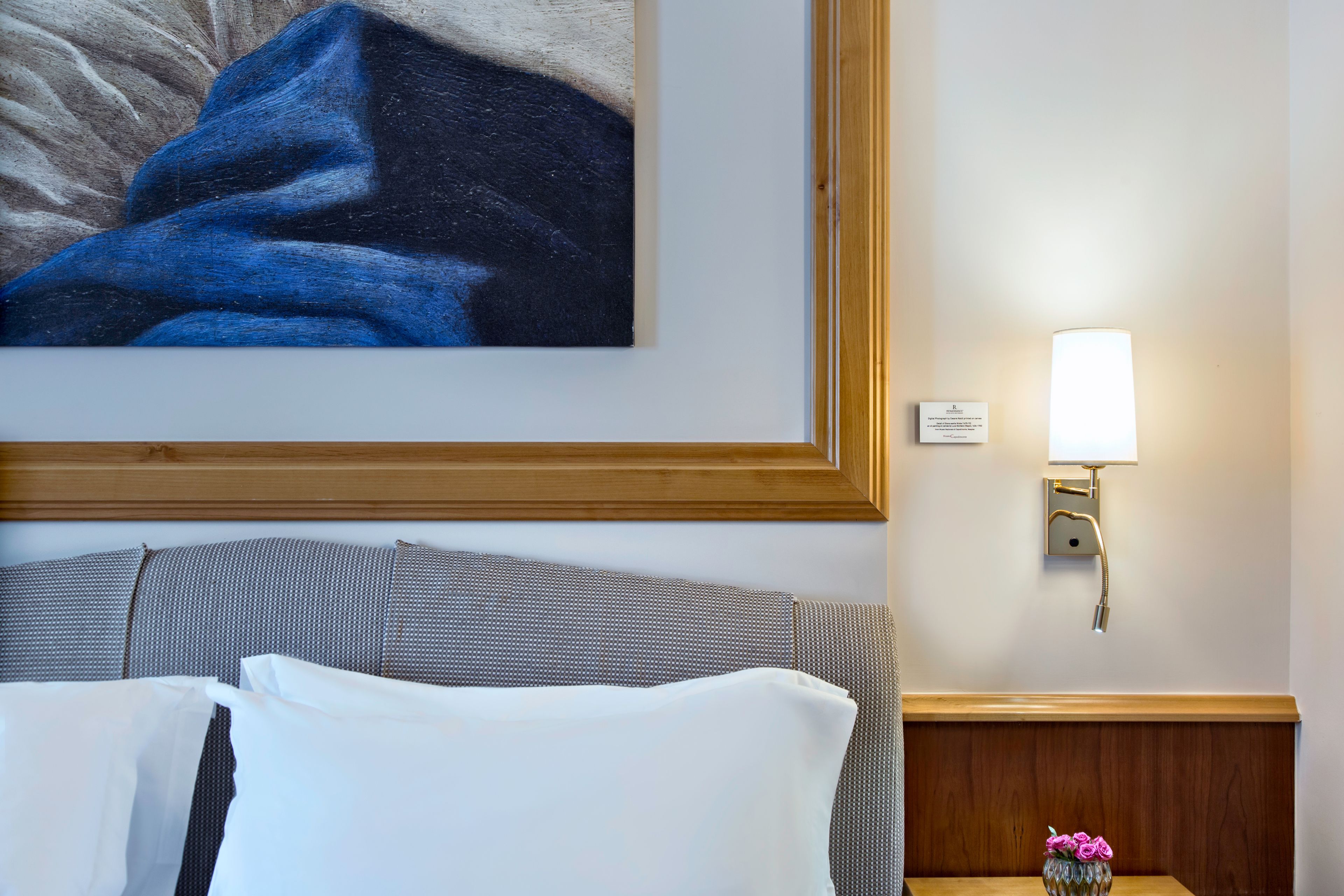 A hotel room with a bed, a nightstand with a lamp, a framed artwork on the wall, and a wooden headboard.