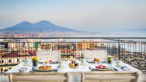 A dining table set on a balcony with a cityscape and mountains in the background. - Renaissance Naples Hotel Mediterraneo (Naples)