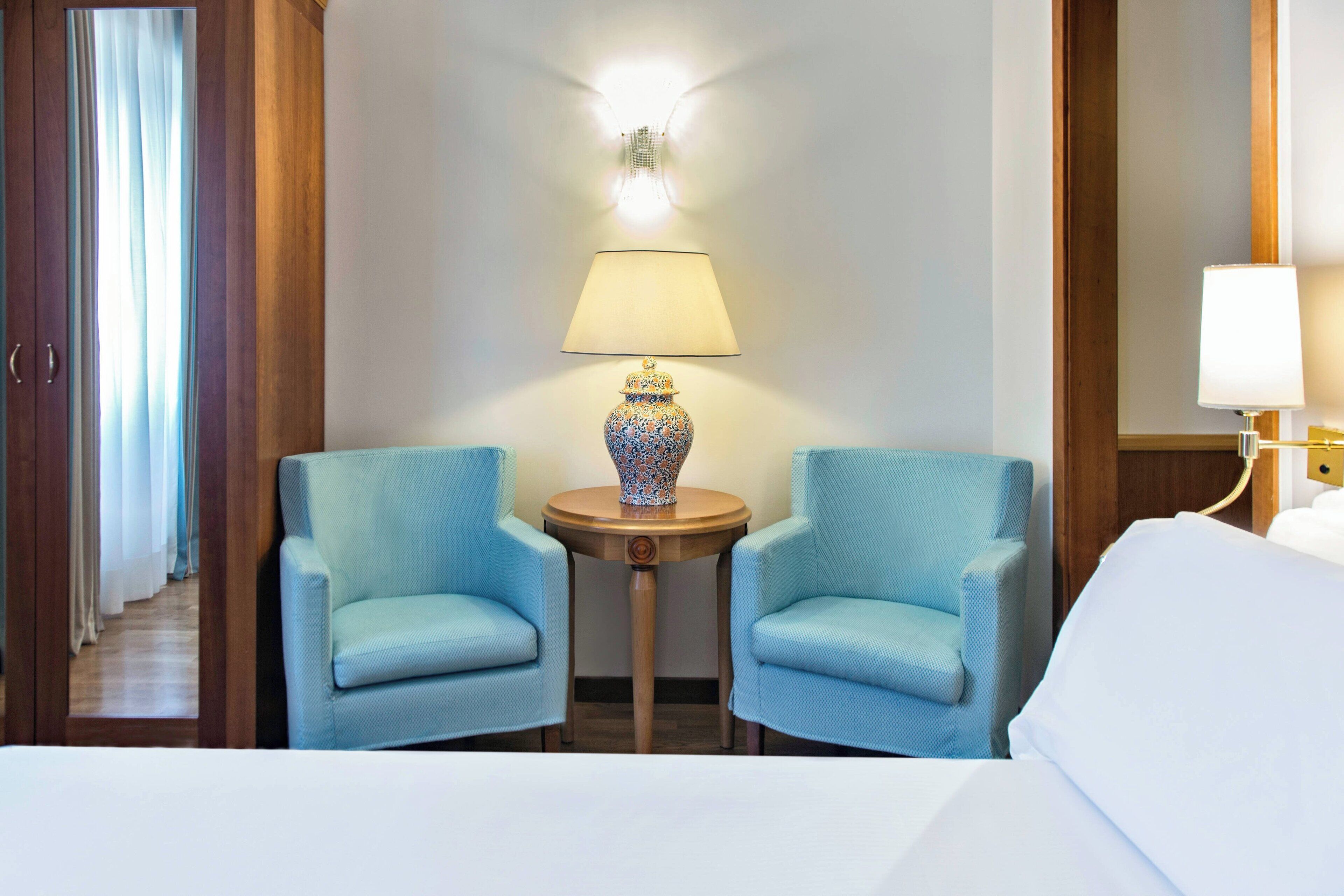 A hotel room with two light blue armchairs, a small round table with a decorative vase, a bed with white linens, and a wall-mounted lamp.