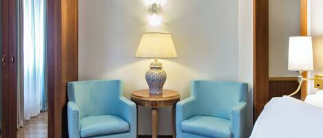 A hotel room with two light blue armchairs, a small round table with a decorative vase, a bed with white linens, and a wall-mounted lamp.